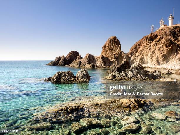 lighthouse on the sea a sunny day on the beach and rocky coast of the cabo de gata with formations of volcanic rock. cabo de gata - nijar natural park, sirens reef, beach, biosphere reserve, almeria,  andalusia, spain - lighthouse reef - fotografias e filmes do acervo