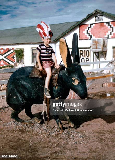 Boy wearing a souvenir Indian headdress sits on a giant cement rabbit at the Jack Rabbit Trading Post on Route 66 near Joseph City, Arizona. The...