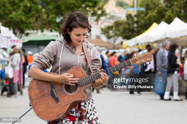 a smiling young woman in casual dress playing a guitar at a farmer's market. - busker stockfoto's en -beelden
