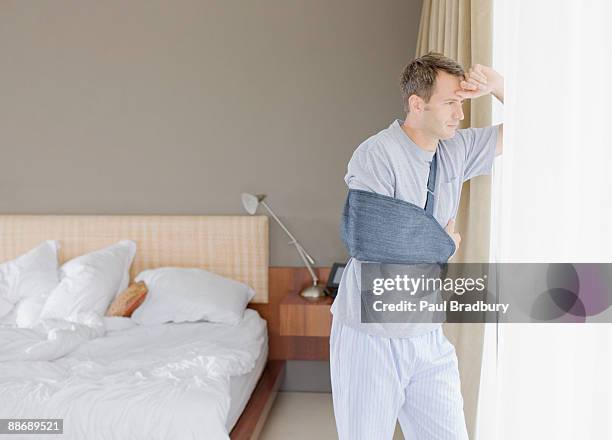 hombre con brazo fracturado mirando por la ventana - cabestrillo-de-brazo fotografías e imágenes de stock