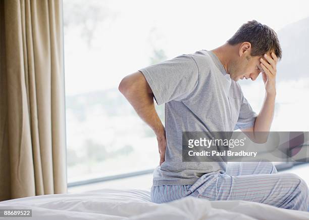 uomo con mal di schiena seduta sul letto e mal di testa - dorsale foto e immagini stock
