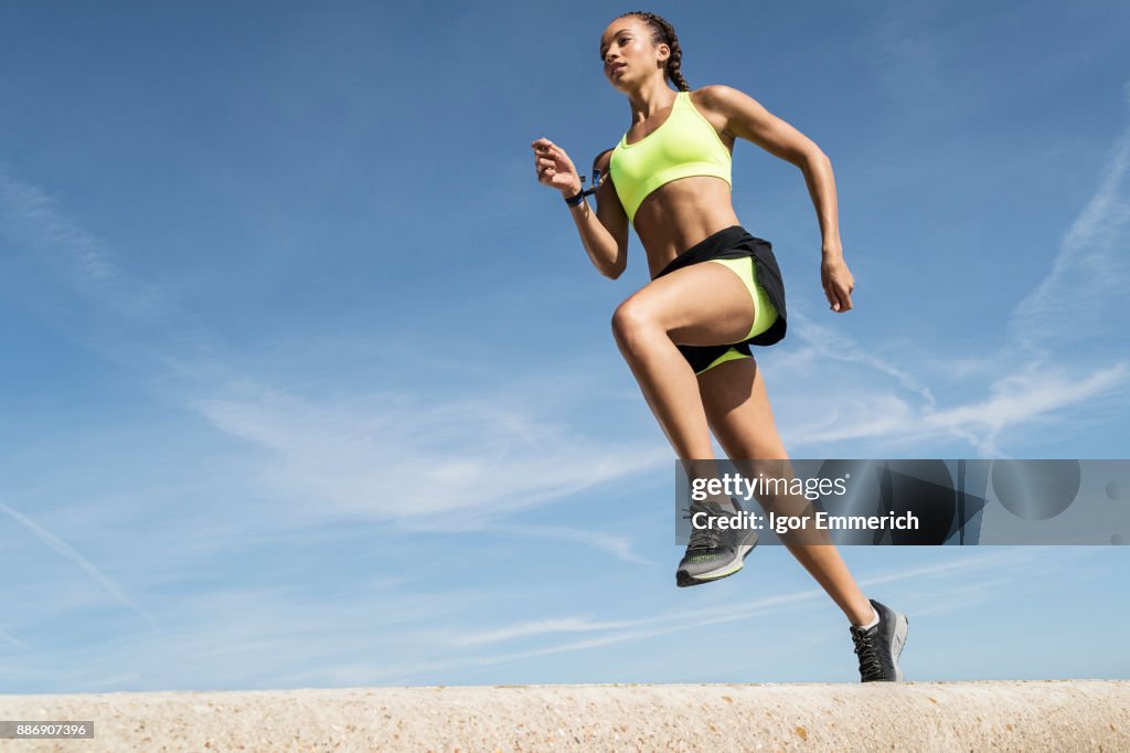 Low angle view of young female runner running along sea wall against blue sky