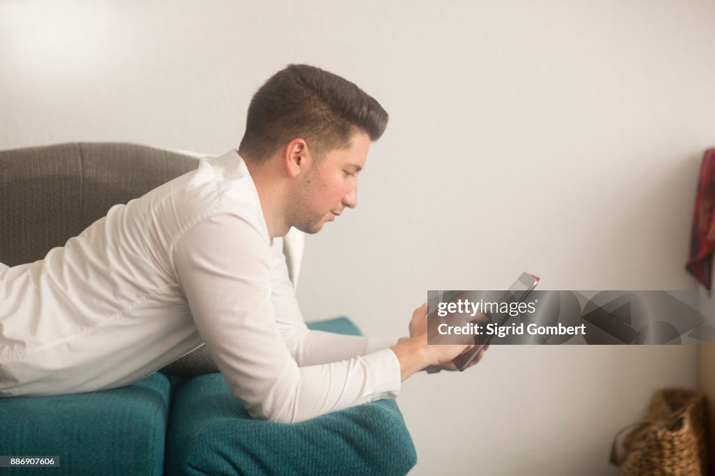 Young man working from home, lying on sofa using digital tablet touchscreen