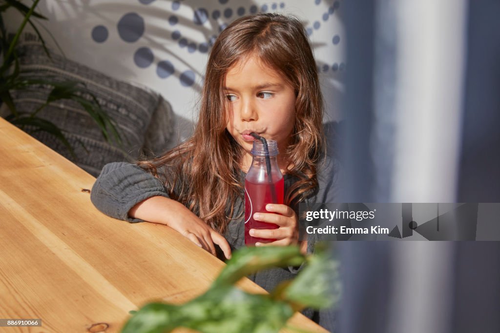 Young girl sitting at table, drinking juice from bottle using straw