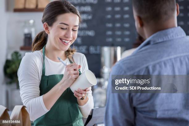 vrolijke coffeeshop barista schrijft volgorde op kop - busy coffee shop stockfoto's en -beelden