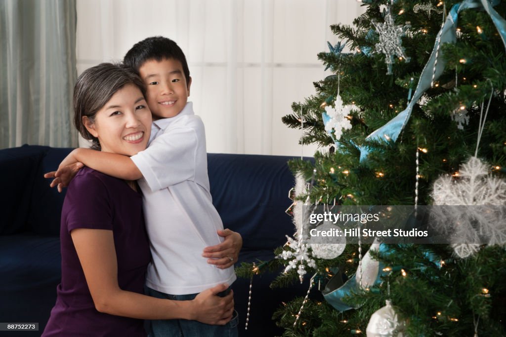 Asian mother hugging son next to Christmas tree