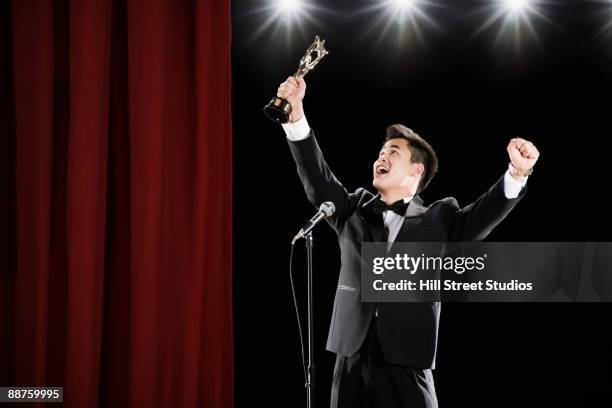 asian man in tuxedo holding trophy overhead at microphone - discurso de agradecimiento fotografías e imágenes de stock