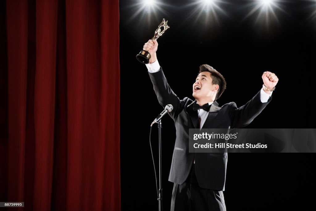 Asian man in tuxedo holding trophy overhead at microphone