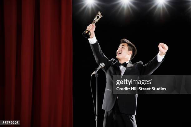 asian man in tuxedo holding trophy overhead at microphone - acceptance speech stock pictures, royalty-free photos & images