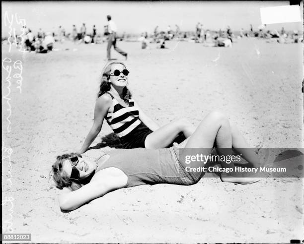 Informal full-length portrait of Elsie Luto and Betty Blee wearing bathing suits and sunglasses, posing on the sand at Wilson Avenue bathing beach in...