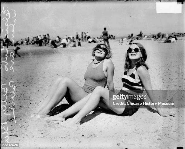 Informal full-length portrait of Elsie Luto and Betty Blee wearing bathing suits and sunglasses and looking up, sitting on the sand at Wilson Avenue...