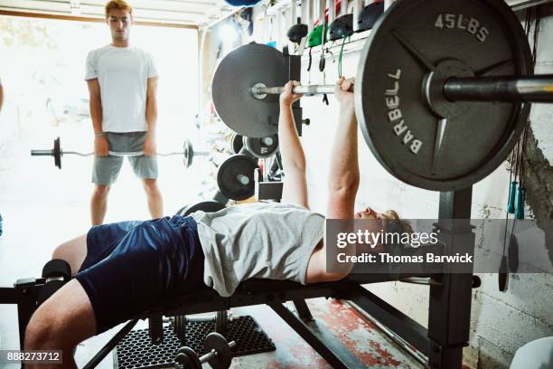 young man doing bench presses while working out with friends in gym in garage - workout bench stock pictures, royalty-free photos & images