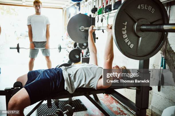 young man doing bench presses while working out with friends in gym in garage - aparelho de musculação imagens e fotografias de stock