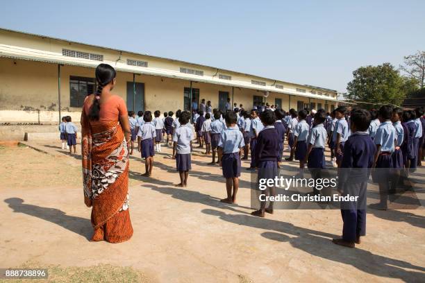 Karon, India Several school boys and girls are standing in line while a female teacher in a orange sari is watching them.