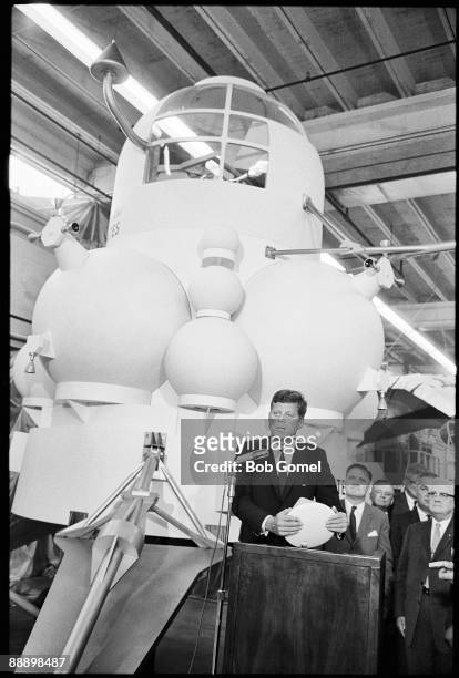American President John F. Kennedy speaks from a lecturn in front of a full-size model of the projected design for a lunar landing module during a...