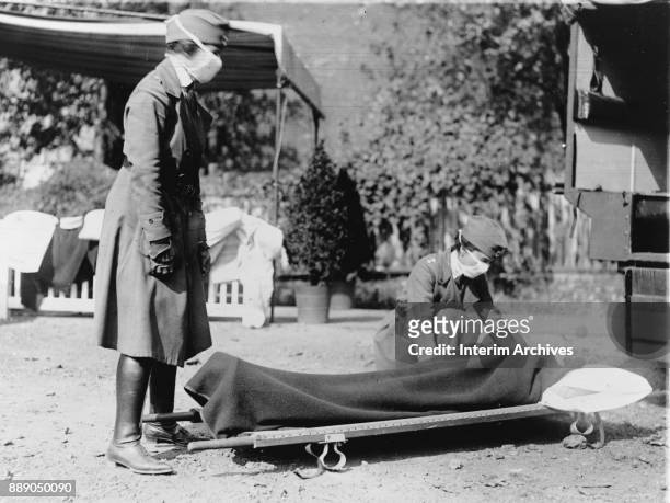During the influenza epidemic, members of the Red Cross Motor Corps, both in uniform with cloth face masks, beside a stretcher on the ground,...