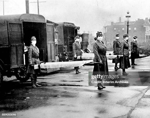 During the influenza epidemic, members of the Red Cross Motor Corps, all in uniform with cloth face masks, pose for a portrait with stretchers behind...