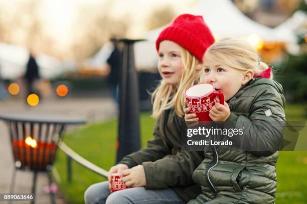 two blonde child sisters drinking hot chocolate at winter european christmas market - family netherlands stock pictures, royalty-free photos & images