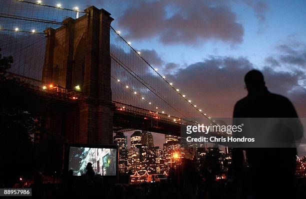 Screen shows a presentation to an audience of hundreds sitting in the grass of Brooklyn Bridge Park across from the Manhattan skyline during the...