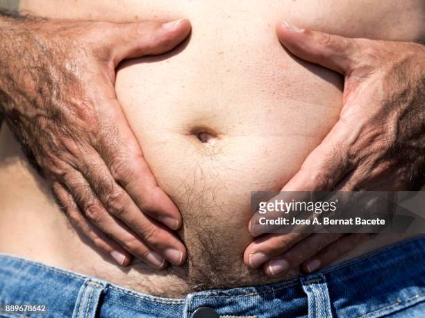 a close-up image of a men mature with  belly fat pinching itself, white-skinned, with jeans shorts in the light of the sun in the beach - belly button stock pictures, royalty-free photos & images