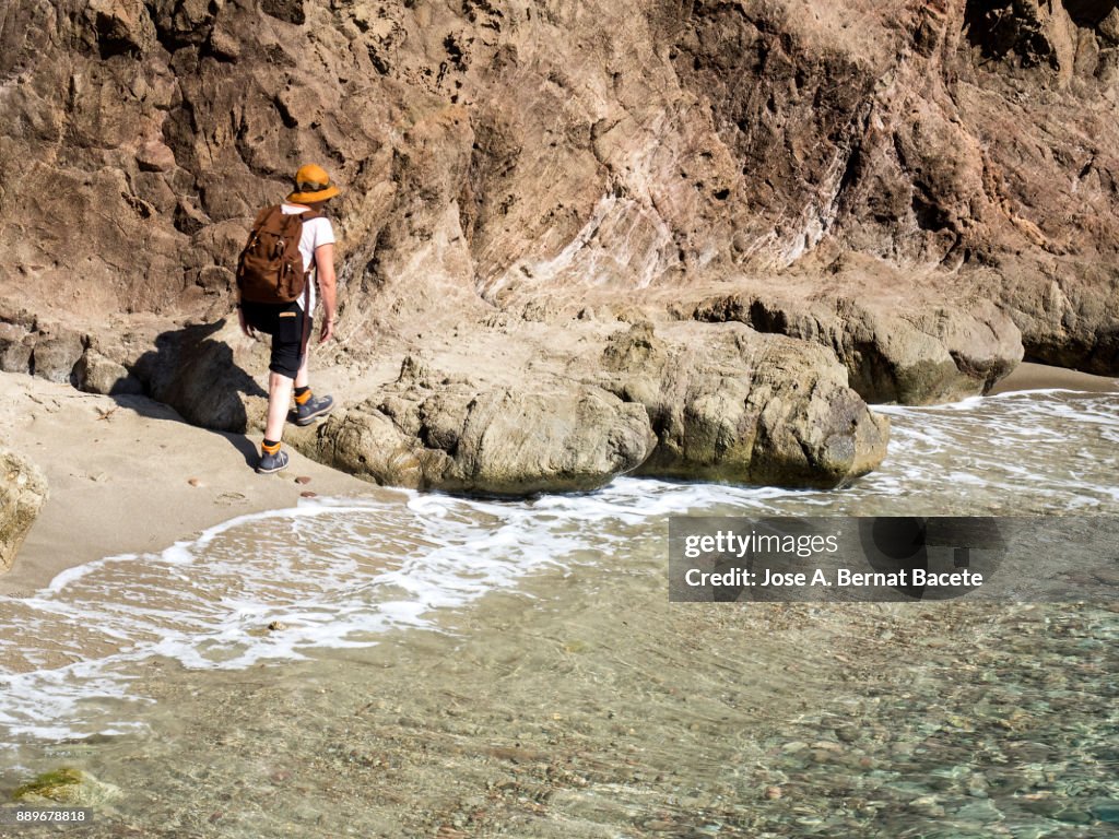 Woman walking along the shore of a beach of sand and rocks of excursion, with a rucksack and a hat. Sirens Reef, Cabo de Gata - Nijar Natural Park, Almeria,  Andalusia, Spain.