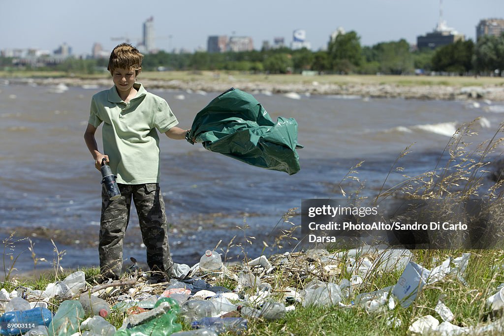 Boy picking up litter on shore, holding garbage bag and magnetic device