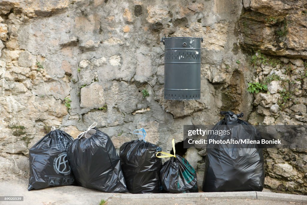 Several bags of garbage lined up along stone wall beneath mounted trash can