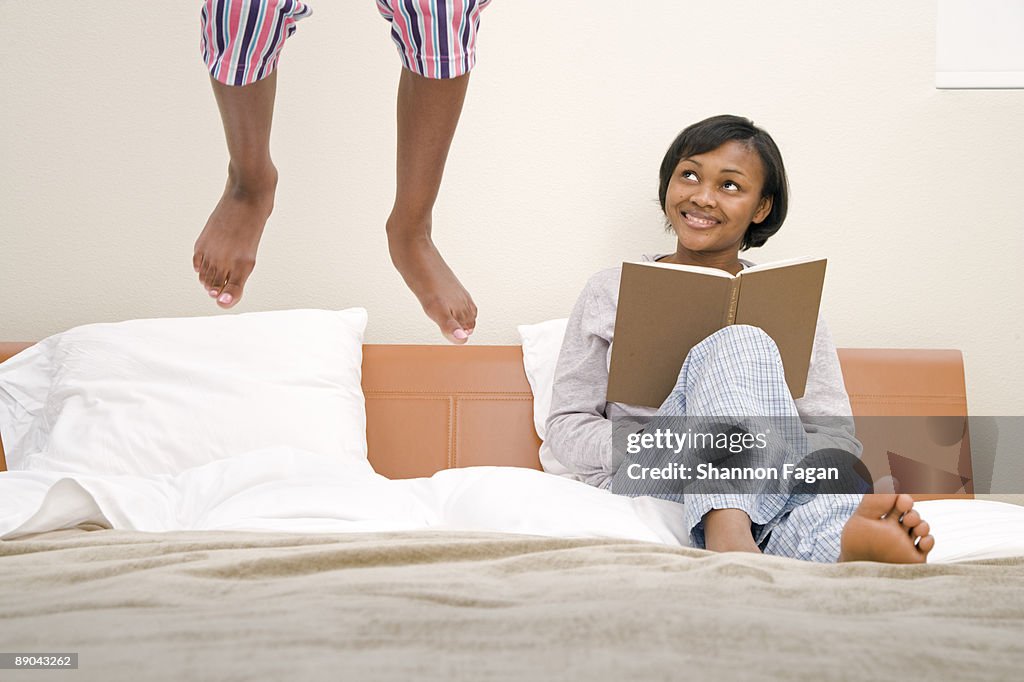 Young Girl Jumping on Bed with Mother Reading Book