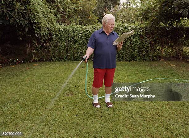 mature man reading newspaper while watering lawn - watering lawn stock pictures, royalty-free photos & images