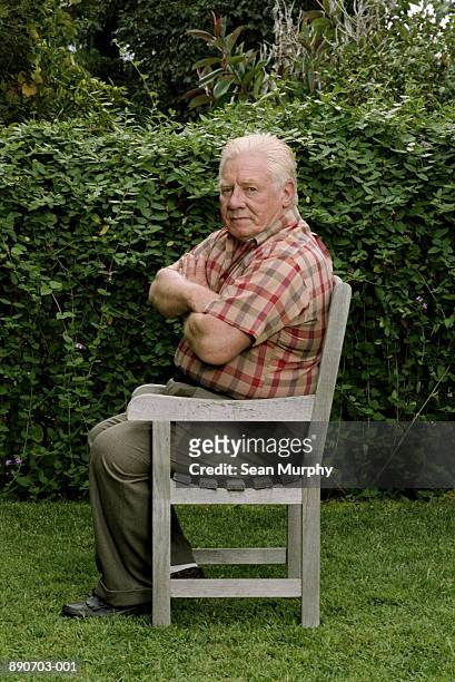 mature man sitting on chair in backyard, arms crossed - grumpy stock-fotos und bilder