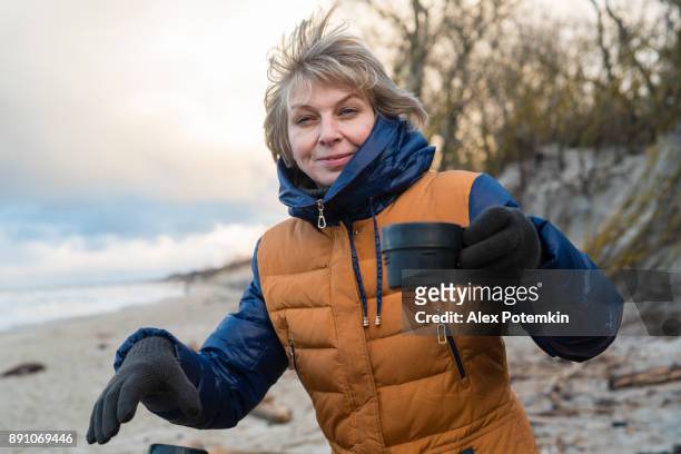 la femme mature séduisante actif âgé de 50 ans à boire le thé chaud de thermos à la plage de la mer baltique froide d’hiver - 50 54 years photos et images de collection