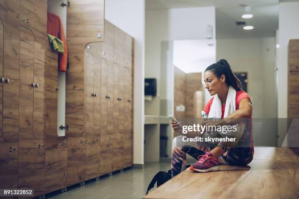young athletic woman sitting on a bench in dressing room and using smart phone. - sports bench stock pictures, royalty-free photos & images