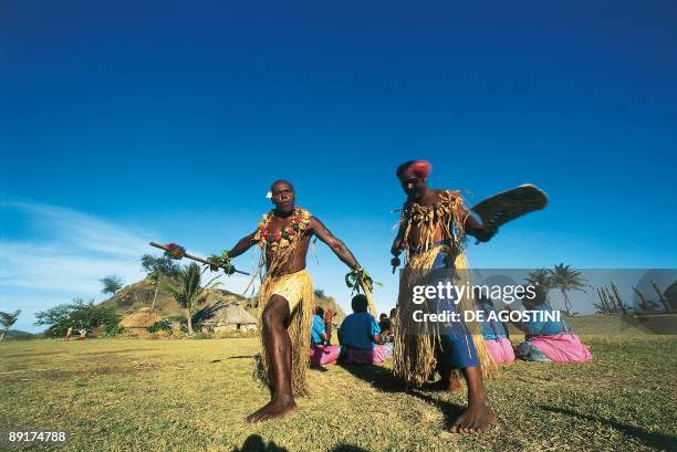 Two mature men performing a tribal dance with group of women sitting behind them, Yasawa Islands, Fiji