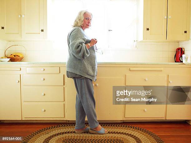 mature woman standing by kitchen sink holding glasses - nosy woman stock pictures, royalty-free photos & images