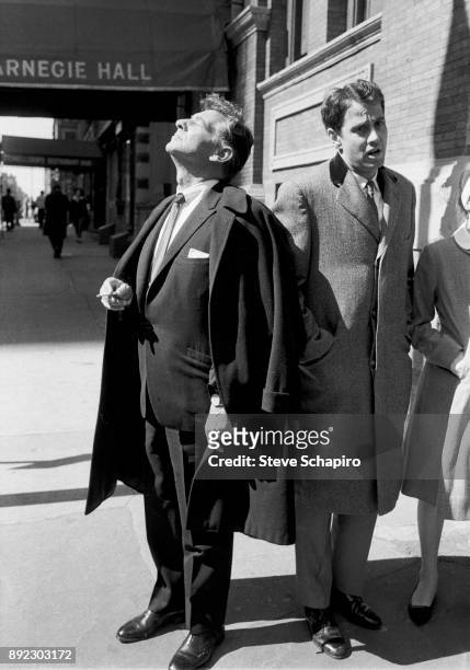 Cigarette in his band, American composer, musician, and conductor Leonard Bernstein stands on the sidewalk outside Carnegie Hall and looks up, eyes...