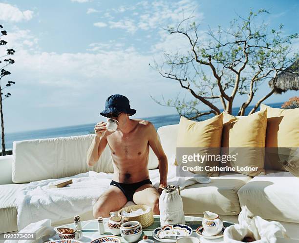 man wearing swimsuit sitting on outdoor couch, drinking coffee - young men in speedos 個照片及圖片檔