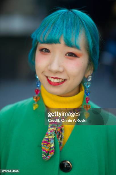 College student and performer Miochin, with dyed blue hair, bright green wool coat, yellow turtleneck sweater, floral print shirt, rainbow scarf,...