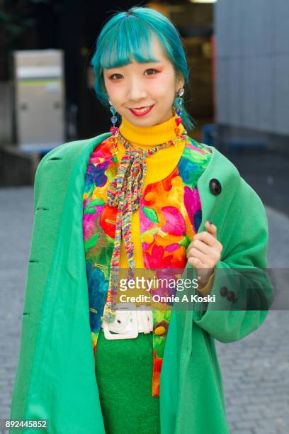 College student and performer Miochin, with dyed blue hair, bright green wool coat, yellow turtleneck sweater, floral print shirt, rainbow scarf,...