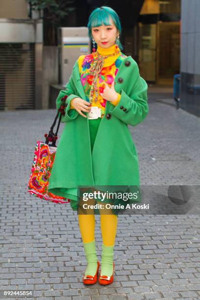 College student and performer Miochin, with dyed blue hair, bright green wool coat, yellow turtleneck sweater, floral print shirt, rainbow scarf,...