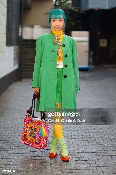 College student and performer Miochin, with dyed blue hair, bright green wool coat, yellow turtleneck sweater, floral print shirt, rainbow scarf,...