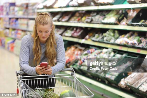 woman in supermarket with trolley, looking at phone - push cart stock pictures, royalty-free photos & images
