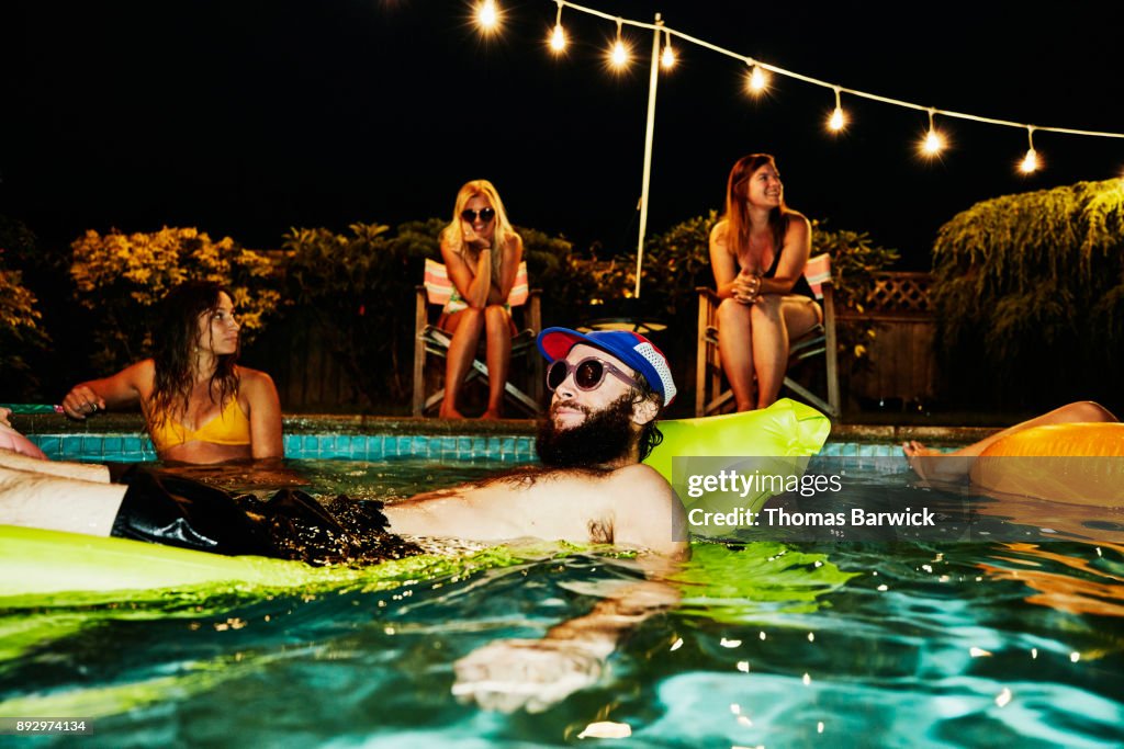 Smiling man wearing sunglasses and floating on air mattress during backyard pool party with friends on summer evening