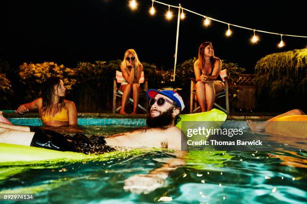 smiling man wearing sunglasses and floating on air mattress during backyard pool party with friends on summer evening - pool party stock pictures, royalty-free photos & images