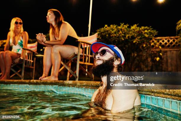 smiling man hanging out in pool during backyard party with friends on summer evening - pool party night stock pictures, royalty-free photos & images