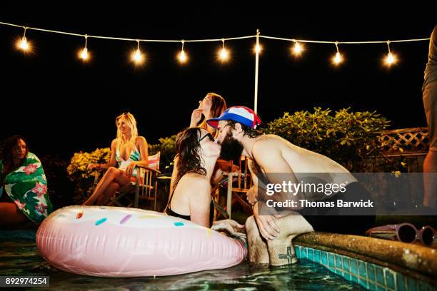 couple kissing while hanging out in pool during backyard party with friends on summer evening - quirky kissing foto e immagini stock