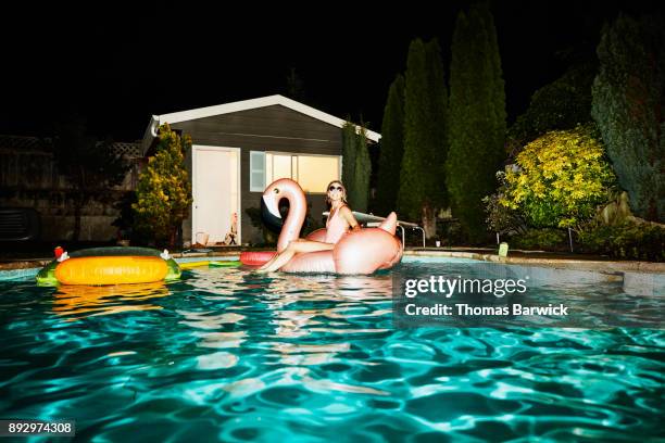 smiling woman wearing sunglasses sitting on inflatable pool toy during backyard pool party on summer evening - pool party night stock pictures, royalty-free photos & images