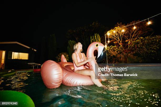 laughing woman riding inflatable pool toy during party with friends on summer evening - pool party night stock pictures, royalty-free photos & images