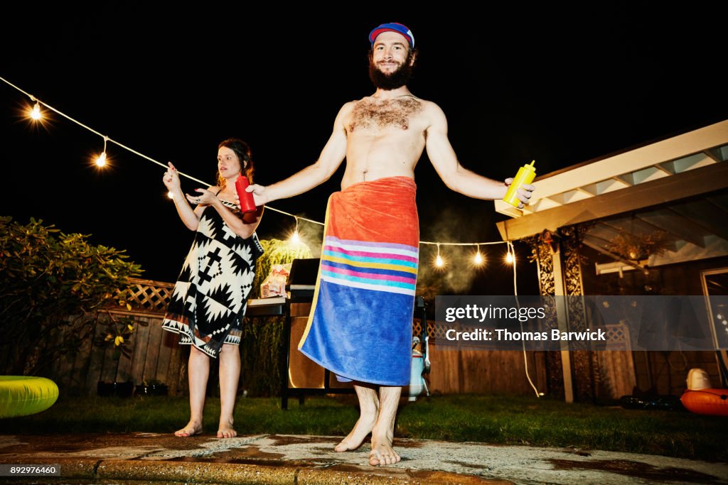 Smiling man wrapped in towel holding ketchup and mustard while grilling for friends in backyard during party on summer evening