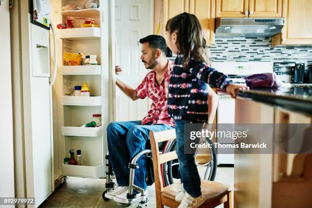 father in wheelchair looking in refrigerator in kitchen while daughter stands on chair watching - disabilitycollection ストックフォトと画像