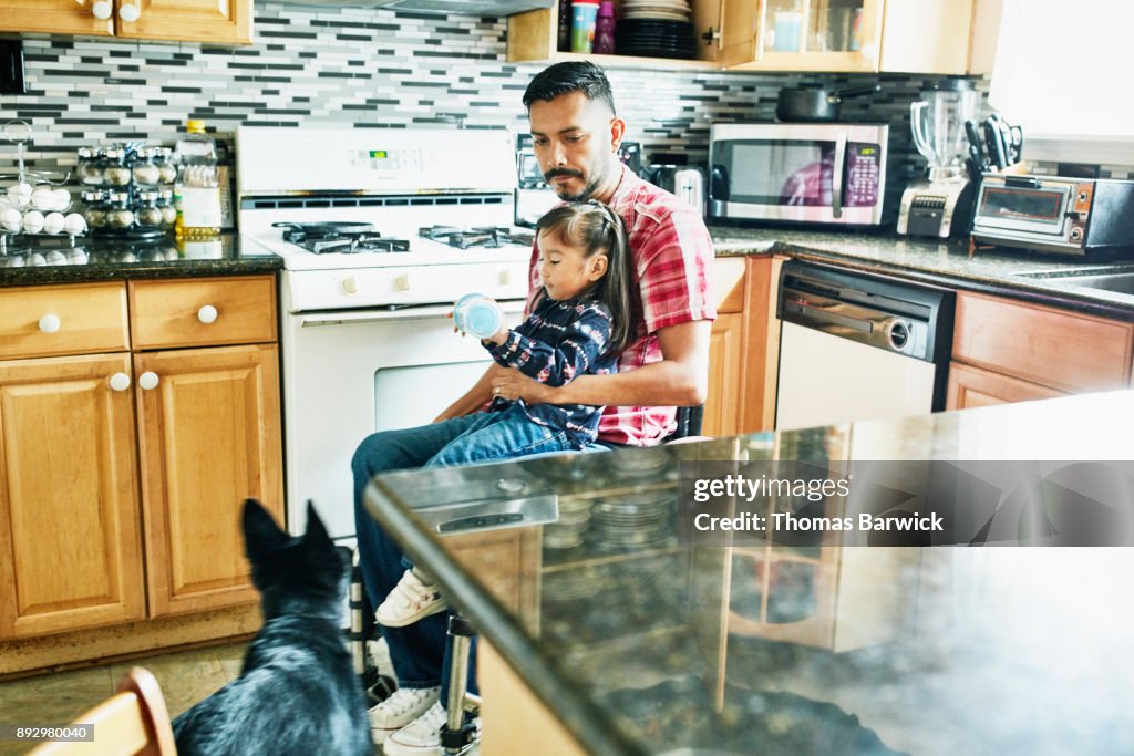 Father in wheelchair holding young daughter in lap while preparing snack in kitchen